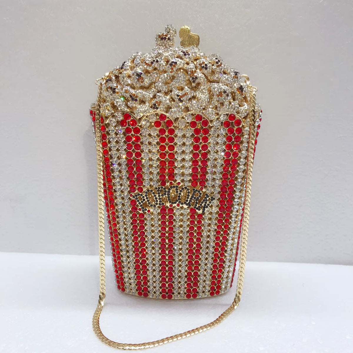 A popcorn-shaped evening bag covered in gold and red rhinestones, with a gold chain strap and a small gold heart on top, photographed against a plain light background.