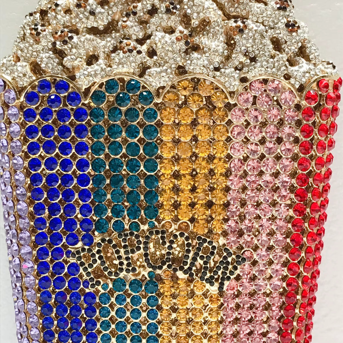 A close-up of a highly embellished, rhinestone-covered popcorn bucket-shaped accessory, featuring vertical columns of blue, teal, gold, pink, and red crystals, with a leopard-print pattern on the top.
